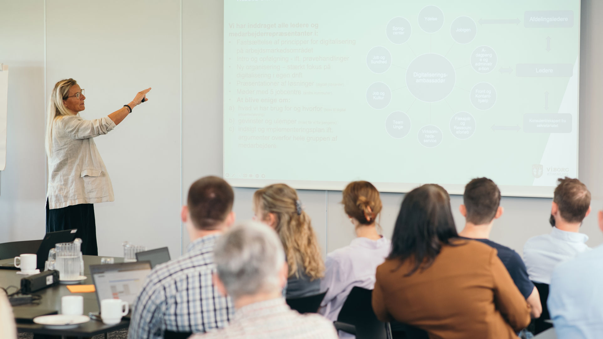 En gruppe mennesker til et møde, hvor en oplægsholder viser slides.