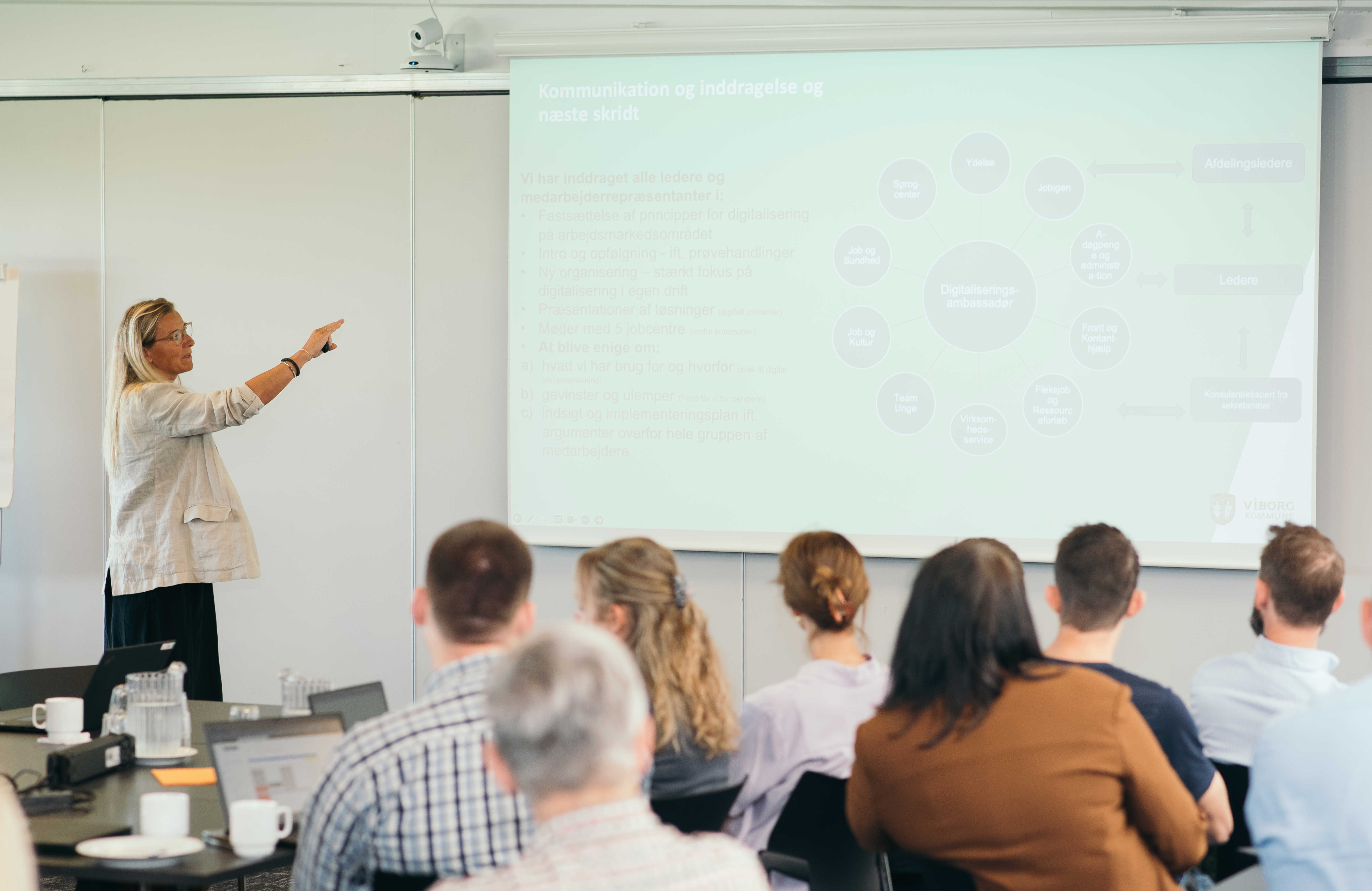 En gruppe mennesker til et møde, hvor en oplægsholder viser slides.
